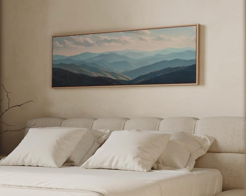 May include: A framed landscape painting of layered blue mountains under a cloudy sky hangs above a bed with white pillows and a cream-colored headboard. The artwork is rectangular with a light wood frame.