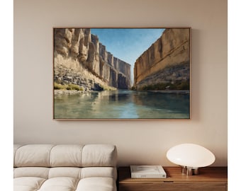May include: A framed landscape painting shows a canyon with high rock formations flanking a river. The artwork uses tan, brown, and blue tones, with a clear blue sky. The painting is displayed above a beige sofa and a wooden bedside table.