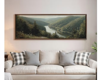 Könnte beinhalten: Gerahmtes Landschaftsgemälde, das einen Fluss zeigt, der sich durch üppige, grüne Hügel schlängelt. Das Kunstwerk hängt über einem cremefarbenen Sofa mit grauen und karierten Kissen. Der Rahmen ist aus dunklem Holz.