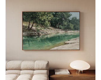 May include: A framed landscape painting shows a tranquil river scene. The water is a vibrant turquoise, reflecting the lush green trees along the banks. The artwork is displayed above a beige sofa and a wooden bedside table with a modern lamp.