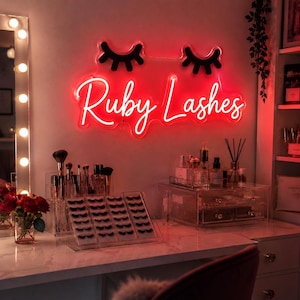 Könnte beinhalten: Ein Schminktisch-Setup mit einem roten Neon-Schild mit der Aufschrift "Ruby Lashes" über einer Marmor-Arbeitsplatte. Auf der Arbeitsplatte befinden sich Make-up-Pinsel, falsche Wimpern und verschiedene Schönheitsprodukte. Links ist ein beleuchteter Spiegel.