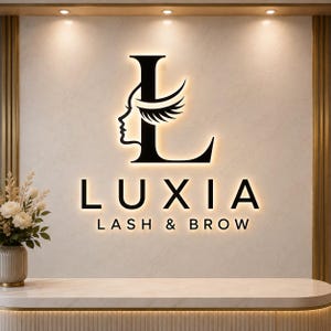 Könnte beinhalten: Ein Wandschild mit dem schwarzen Schriftzug LUXIA LASH & BROW, bei dem ein großer Buchstabe L mit der Profilsilhouette eines Gesichts und Wimpern kombiniert ist. Das Schild ist vor einer hellen Marmorwand warm hinterleuchtet.