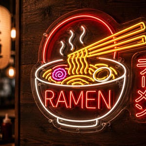 Könnte beinhalten: Ein Neonschild in Form einer Ramen-Schüssel mit Essstäbchen, Nudeln und dem Wort "RAMEN" in Rot. Das Schild ist weiß und rot umrandet, mit einer japanischen Laterne im Hintergrund.