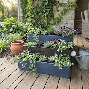 May include: A three-tiered wooden planter box with a dark blue finish, featuring various green succulents, trailing ivy, and purple and white flowers planted in each level. The rectangular structure is positioned on a wooden deck surface.