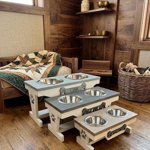 May include: Three tiered pet feeding stations with white wooden bases featuring paw print cutouts and grey stone-like tops. Each station holds two stainless steel bowls and includes a bone-shaped nameplate labeled Tank, Rosco, or Dalton.