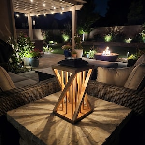 Peut inclure: Une lanterne d'ext&eacute;rieur en bois au design g&eacute;om&eacute;trique est pos&eacute;e sur une table en pierre. La lumi&egrave;re de la lanterne projette un motif en &eacute;toile sur la surface de la table. L'arri&egrave;re-plan pr&eacute;sente un cadre de patio avec un foyer et des guirlandes lumineuses.