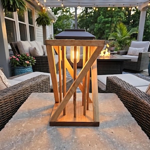 Peut inclure: Une lanterne en bois avec un dessus en m&eacute;tal fonc&eacute; et une ampoule &agrave; l'int&eacute;rieur. La lanterne a un design g&eacute;om&eacute;trique avec des supports en bois verticaux et inclin&eacute;s. Elle repose sur une surface en pierre, avec des si&egrave;ges ext&eacute;rieurs et des guirlandes lumineuses en arri&egrave;re-plan.