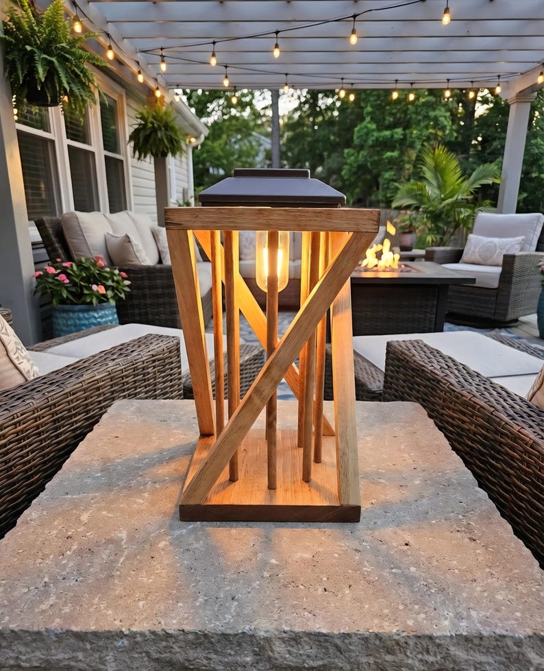 Peut inclure: Une lanterne en bois avec un dessus en m&eacute;tal fonc&eacute; et une ampoule &agrave; l'int&eacute;rieur. La lanterne a un design g&eacute;om&eacute;trique avec des supports en bois verticaux et inclin&eacute;s. Elle repose sur une surface en pierre, avec des si&egrave;ges ext&eacute;rieurs et des guirlandes lumineuses en arri&egrave;re-plan.
