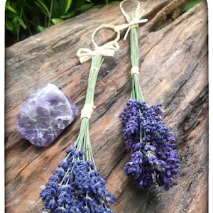 May include: Two bunches of dried lavender with purple flowers and green stems, tied with raffia bows. A raw amethyst crystal sits beside the lavender on a weathered wooden surface. The image has a natural, rustic aesthetic.