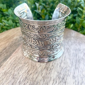 May include: Silver cuff bracelet with detailed embossed patterns. The wide bracelet has intricate designs and a polished finish. The open design allows for easy wear. The bracelet is displayed on a wooden surface with green foliage in the background.