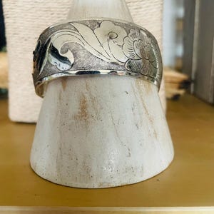 May include: A silver cuff bracelet with an engraved floral pattern. The wide band displays detailed carvings of flowers and leaves. The bracelet is presented on a white, cone-shaped stand, highlighting its intricate craftsmanship.