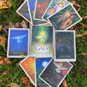 May include: A selection of oracle cards with diverse imagery and text, including "Gaia Oracle" and "Rising Above." The cards display illustrations of doves, trees, and spiritual figures, set on a bed of green grass and fallen leaves.