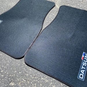 May include: Two black car floor mats with the word "DATSUN" in a white, red, and blue logo. The mats are made of a dark gray, textured material and have a black border.
