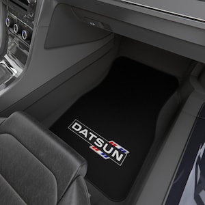 May include: Black car floor mat with the Datsun logo in white, red, and blue. The mat is inside a car, with the dashboard and seat visible in the background. The Datsun logo is in a rectangular box.