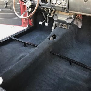 May include: Interior view of a classic car, showcasing the black carpeted floor and dashboard. The steering wheel, pedals, and gear shift are visible, along with the red painted door panel. The car's interior is in good condition.