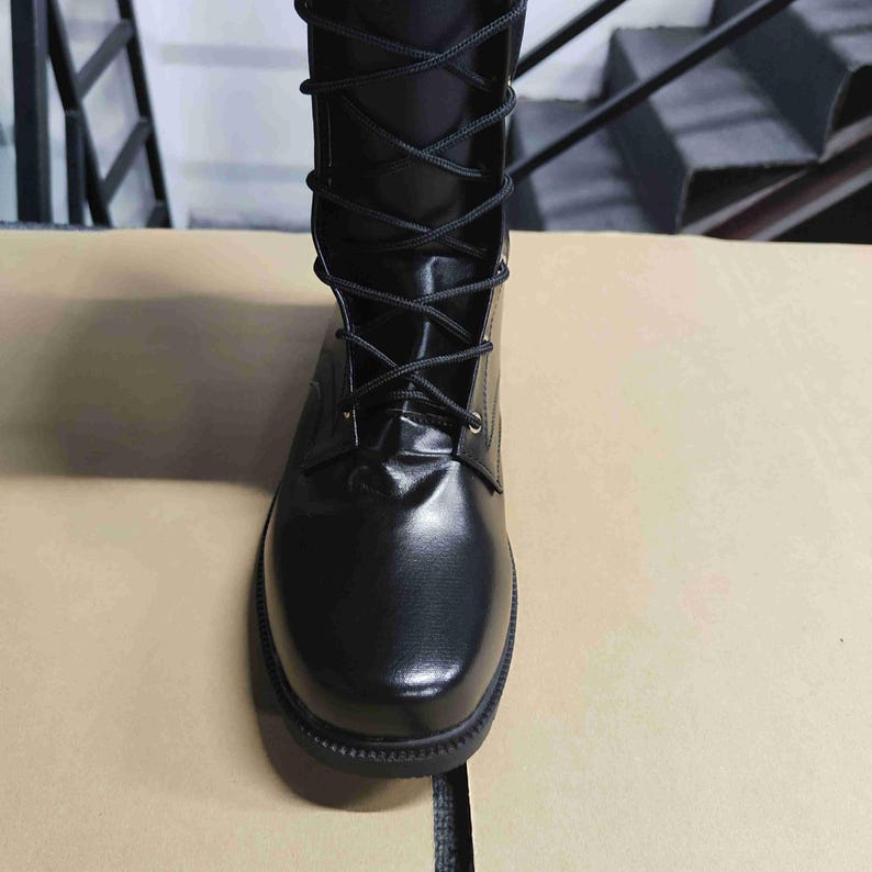 Puede incluir: Una bota negra de media ca&ntilde;a con un acabado liso y brillante y puntera redondeada. La bota presenta un cierre frontal con cordones negros finos a trav&eacute;s de ojales met&aacute;licos, que se extiende desde la base del pie hasta la parte superior.