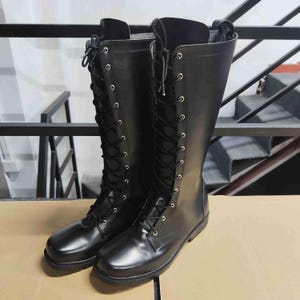 Puede incluir: Un par de botas negras hasta la rodilla con acabado liso, punteras redondeadas y cierre frontal de cordones con ojales met&aacute;licos plateados. Las botas tienen una suela negra s&oacute;lida y una ca&ntilde;a estructurada que llega hasta la parte inferior de la rodilla.