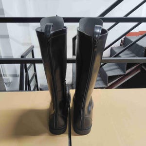 Puede incluir: Un par de botas altas negras con acabado liso y una cremallera vertical completa en la parte posterior de cada ca&ntilde;a. Las botas tienen un dise&ntilde;o estructurado con una leng&uuml;eta en la parte superior de la cremallera y una suela plana, vista trasera.