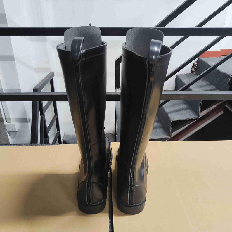 Puede incluir: Un par de botas altas negras con acabado liso y una cremallera vertical completa en la parte posterior de cada ca&ntilde;a. Las botas tienen un dise&ntilde;o estructurado con una leng&uuml;eta en la parte superior de la cremallera y una suela plana, vista trasera.