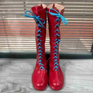 May include: A pair of knee-high red boots with bright blue laces. The boots have a glossy finish and a round toe. The laces are tied in a crisscross pattern up the front of the boots. The boots are displayed against a neutral background.