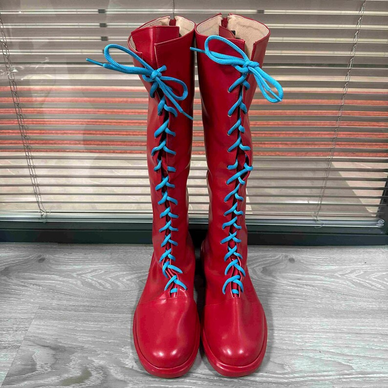 May include: A pair of knee-high red boots with bright blue laces. The boots have a glossy finish and a round toe. The laces are tied in a crisscross pattern up the front of the boots. The boots are displayed against a neutral background.