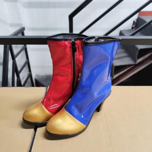 May include: Pair of shiny, knee-high boots. One boot is red with a black zip, the other is blue. Both boots have gold-coloured toes and black heels. The boots are made of a glossy material.