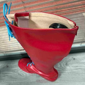 May include: A tall, red leather boot with bright blue laces. The boot has a rounded toe and a high shaft, with a contrasting beige interior. A black object is visible inside the boot.