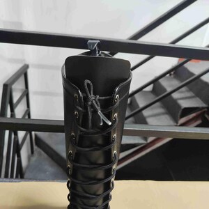 Puede incluir: Una bota alta negra con acabado mate liso, ca&ntilde;a alta y cierre frontal de cordones negros que pasan por ojales met&aacute;licos de tono plateado. La bota est&aacute; sujeta en la parte superior con un gancho de suspensi&oacute;n de pl&aacute;stico negro.