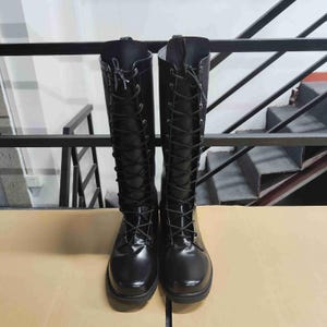 Puede incluir: Un par de botas negras hasta la rodilla con acabado brillante, punta redondeada y cierre frontal completo de cordones negros que pasan por ojales met&aacute;licos plateados, extendi&eacute;ndose desde la punta hasta la parte superior de la ca&ntilde;a.