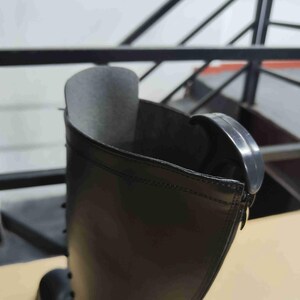 Puede incluir: Una bota negra con una ca&ntilde;a alta de acabado liso y mate. Se observa la abertura superior de la bota, mostrando un forro interior oscuro y una leng&uuml;eta de pl&aacute;stico negro curvada unida al borde exterior cerca de la costura superior de la ca&ntilde;a.