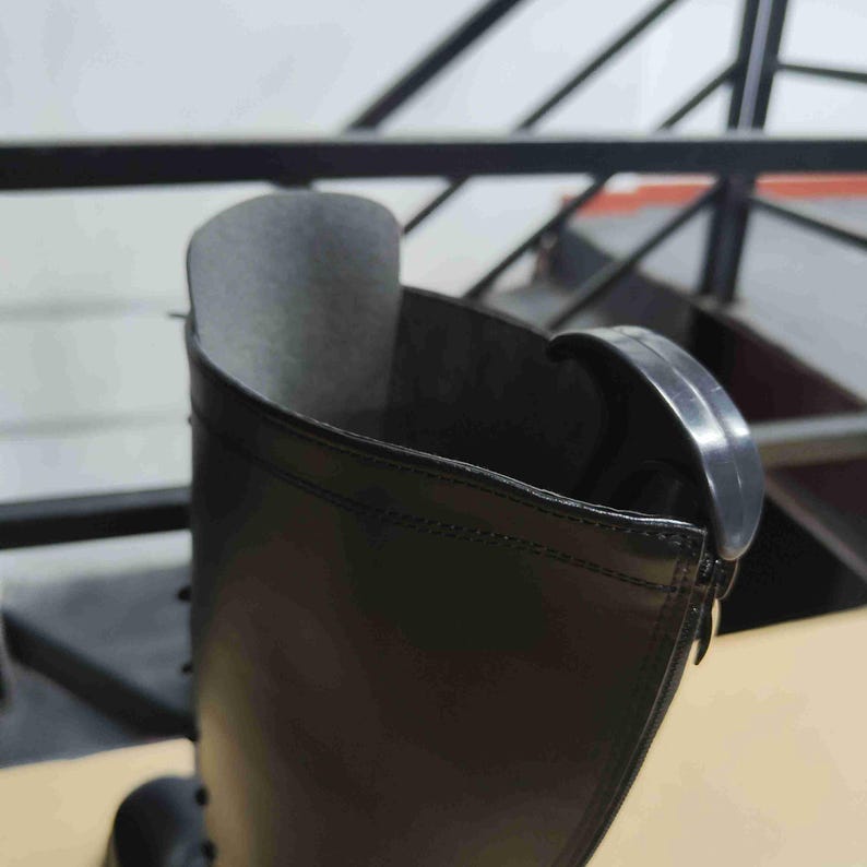 Puede incluir: Una bota negra con una ca&ntilde;a alta de acabado liso y mate. Se observa la abertura superior de la bota, mostrando un forro interior oscuro y una leng&uuml;eta de pl&aacute;stico negro curvada unida al borde exterior cerca de la costura superior de la ca&ntilde;a.
