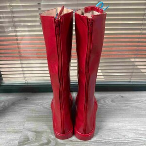 May include: A pair of tall, red boots with zippers running down the back. The boots are made of a shiny material and have a flat sole. The zippers are the same color as the boots. The boots are knee-high.