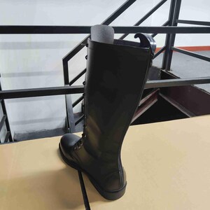 Puede incluir: Una bota alta de color negro con dise&ntilde;o de cordones frontales, ojales met&aacute;licos, punta redondeada y suela gruesa. La bota tiene un acabado liso y una trabilla en la parte superior trasera, con un inserto de espuma gris visible en el interior.