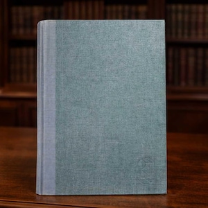 May include: A hardcover book with a teal cover and a light gray spine stands upright on a dark wooden surface. The book has a textured appearance and a subtle embossed design in the lower right corner. The background features a dimly lit room with bookshelves and a lamp.