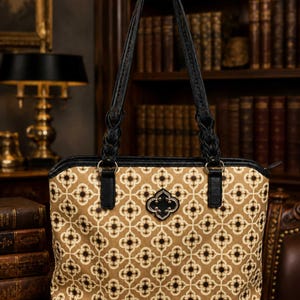 May include: A tan tote bag with a repeating floral pattern in white and dark brown. The bag has black leather handles and trim, with a decorative black metal clasp. The handles are braided. The bag is sitting on a wooden surface.