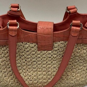 Peut inclure: Un sac fourre-tout en paille tress&eacute;e avec des accents de cuir couleur corail. Le sac pr&eacute;sente un dessus, des poign&eacute;es et un fermoir en cuir corail textur&eacute; crocodile. Des anneaux m&eacute;talliques argent&eacute;s relient les poign&eacute;es au sac.