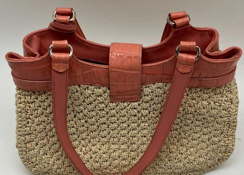 Peut inclure: Un sac fourre-tout en paille tress&eacute;e avec des accents de cuir couleur corail. Le sac pr&eacute;sente un dessus, des poign&eacute;es et un fermoir en cuir corail textur&eacute; crocodile. Des anneaux m&eacute;talliques argent&eacute;s relient les poign&eacute;es au sac.