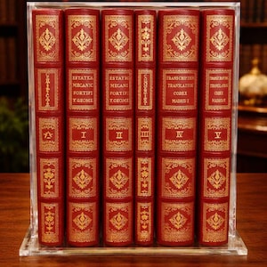 May include: A collection of antique books in a clear display case. The books have red covers with gold embossed decorative patterns and text. The spines display titles and volume numbers. The books are arranged on a wooden surface.