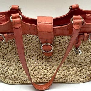 Peut inclure: Un sac fourre-tout en paille tress&eacute;e avec des d&eacute;tails en cuir corail. Le sac pr&eacute;sente un corps en paille tress&eacute;e, des lani&egrave;res en cuir corail et un rabat en cuir corail avec une boucle argent&eacute;e. Il a une garniture argent&eacute;e et un charm en forme de c&oelig;ur.