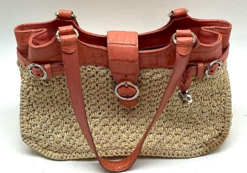Peut inclure: Un sac fourre-tout en paille tress&eacute;e avec des d&eacute;tails en cuir corail. Le sac pr&eacute;sente un corps en paille tress&eacute;e, des lani&egrave;res en cuir corail et un rabat en cuir corail avec une boucle argent&eacute;e. Il a une garniture argent&eacute;e et un charm en forme de c&oelig;ur.