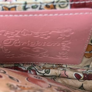 Peut inclure: Gros plan sur l'int&eacute;rieur d'un portefeuille en cuir rose. Le portefeuille pr&eacute;sente le nom de la marque "Brighton" en relief et des d&eacute;tails floraux. La doublure int&eacute;rieure pr&eacute;sente un motif floral dans les tons de rose, de vert et de beige.