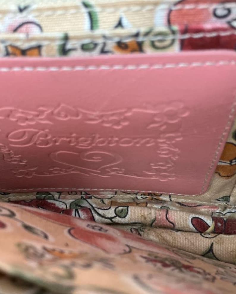 Peut inclure: Gros plan sur l'int&eacute;rieur d'un portefeuille en cuir rose. Le portefeuille pr&eacute;sente le nom de la marque "Brighton" en relief et des d&eacute;tails floraux. La doublure int&eacute;rieure pr&eacute;sente un motif floral dans les tons de rose, de vert et de beige.