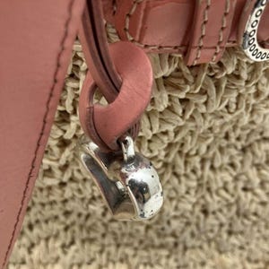 Peut inclure: Gros plan sur un sac &agrave; main en cuir rose avec une base en paille tress&eacute;e. Le sac est orn&eacute; d'un charm argent&eacute; et d'une sangle et d'une boucle en cuir rose. Le design du sac est simple et &eacute;l&eacute;gant.