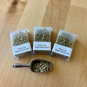 May include: Three clear plastic containers filled with small, beige-colored beads, labeled "Toho 11-369 Beige Lined Crystal." A silver scoop holds more beads. The items are arranged on a light brown wooden surface.