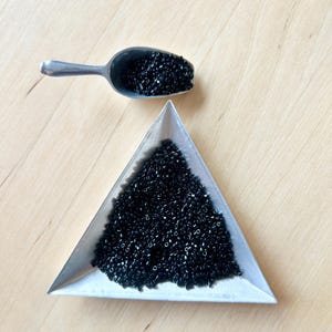 Peut inclure: Vue de dessus d'une cuillère métallique et d'un plateau triangulaire remplis de petites perles noires brillantes. Les objets sont posés sur une surface en bois clair.
