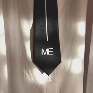 Puede incluir: Corbata negra con una raya vertical blanca y la palabra "ME" impresa en blanco. La corbata cuelga de una camisa blanca abotonada.