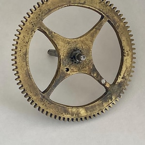 May include: A vintage brass gear with numerous teeth around the outer edge. The gear has a central hub with a small pin and a cross-shaped spoke design. The metal has a slightly tarnished appearance, suggesting age and use. This gear is likely from a clock or other mechanical device.