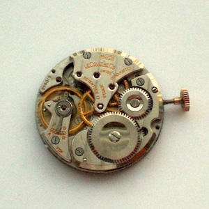Pu&ograve; includere: Primo piano di un movimento di orologio con ingranaggi, viti e la scritta "LeCoultre Co Swiss". Il meccanismo dell'orologio presenta toni oro e argento, con una piccola corona. Il movimento dell'orologio &egrave; contrassegnato "Seventeen 17 Jewels".