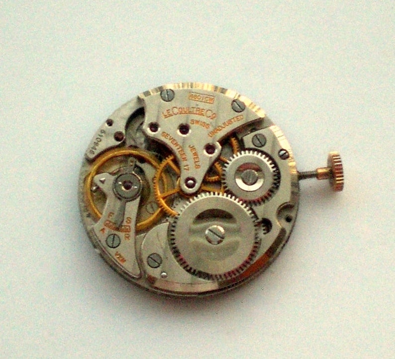 Pu&ograve; includere: Primo piano di un movimento di orologio con ingranaggi, viti e la scritta "LeCoultre Co Swiss". Il meccanismo dell'orologio presenta toni oro e argento, con una piccola corona. Il movimento dell'orologio &egrave; contrassegnato "Seventeen 17 Jewels".