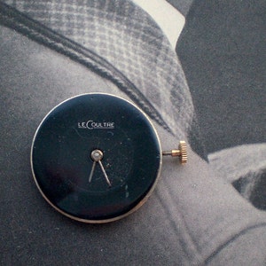 Pu&ograve; includere: Un quadrante nero con il marchio "LE COULTRE" in lettere bianche. Presenta due lancette argentate e una corona dorata. Il quadrante ha un bordo dorato. Il diametro &egrave; di circa 3 cm.